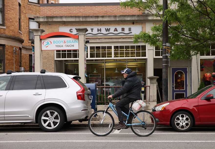 An older man in a baseball cap and parka rides a blue bicycle on a city shopping street, past parked cars and a yoga studio