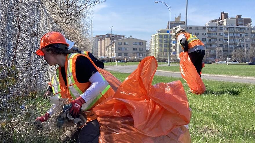 Des travailleurs portant un dossard et tenant des sacs poubelles ramassent des déchets sur une étendue gazonnée en bordure d'une bretelle d'autoroute.