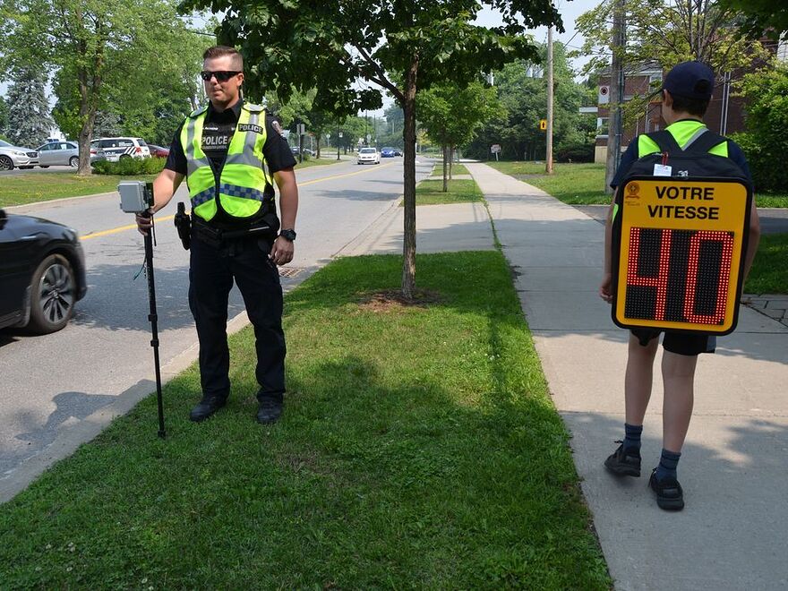 Children in the Montreal area are becoming living speed cameras as a way to get drivers to slow down in school zones. A police officer stands by as a child with a backpack is equipped with a readout showing drivers' speeds, in a Friday, June 30, 2023, handout photo.
