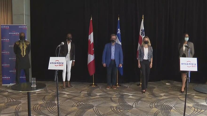 Cinq personnes masquées devant des drapeaux du Canada, du Québec et de Montréal, avec des micros à l'avant-plan.