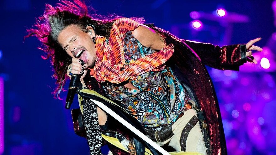 Steven Tyler, qui porte une tenue très colorée, chante au micro sur scène.