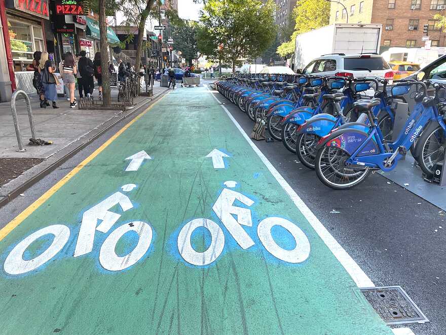 A bike lane painted bright green separates a busy sidewalk with a pizza place and other shops from a bikeshare facility full of blue Citibikes and automotive traffic.