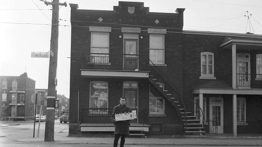 Un homme tient une affiche numérotée devant des maisons de l'avenue Decelles.