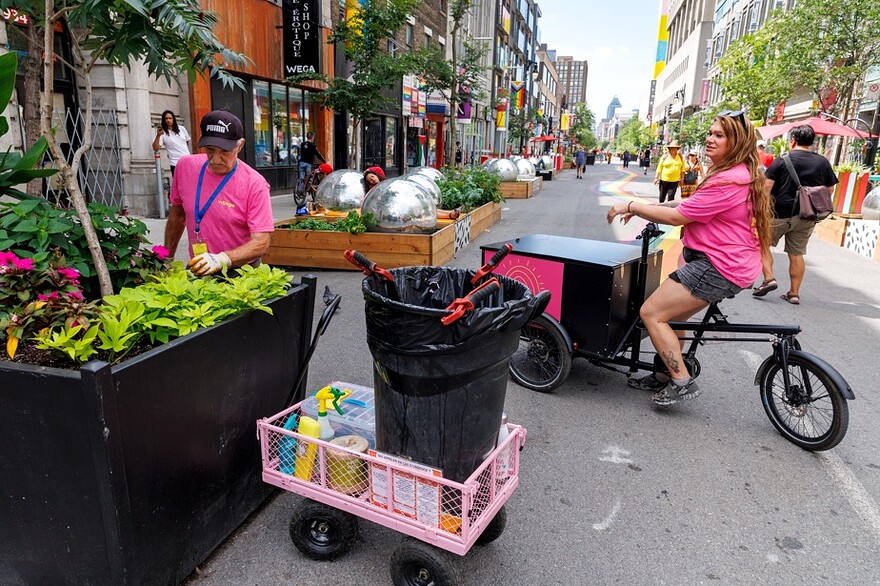 Claude Poirier tire une poubelle dans un chariot rose et Sasha Baga conduit un tricycle électrique dont le coffre avant est rempli de bidons d’eau.