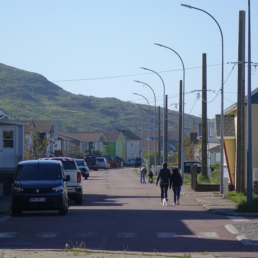 Deux piétons marchent dans la rue d'un village avec un petit mont dans l'arrière-fond.