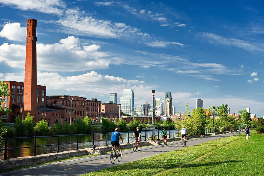 Bicyclists ride on a paved bike path. On one side is an expanse of lawn, and on the other side is a densely built urban area, with tall modern buildings rising in the distance.