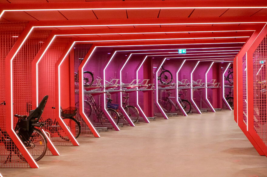 Photos of a futuristic-looking, brightly-lit, colourful bike parking facility and public passage beneath Berlin’s Potsdamer Platz. The red and purple curvilinear space is lined with a variety of vertical and horizontal racks, each separated by a metal grill structure which incorporates LED lighting.