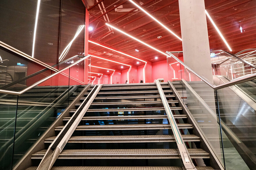Photos of a futuristic-looking, brightly-lit, colourful bike parking facility and public passage beneath Berlin’s Potsdamer Platz. The red and purple curvilinear space is lined with a variety of vertical and horizontal racks, each separated by a metal grill structure which incorporates LED lighting.