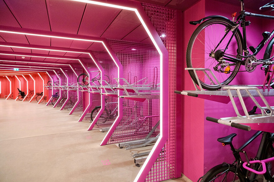 Photos of a futuristic-looking, brightly-lit, colourful bike parking facility and public passage beneath Berlin’s Potsdamer Platz. The red and purple curvilinear space is lined with a variety of vertical and horizontal racks, each separated by a metal grill structure which incorporates LED lighting.
