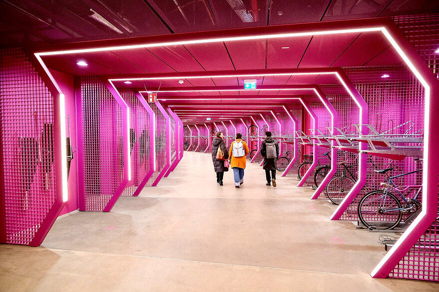 Photos of a futuristic-looking, brightly-lit, colourful bike parking facility and public passage beneath Berlin’s Potsdamer Platz. The red and purple curvilinear space is lined with a variety of vertical and horizontal racks, each separated by a metal grill structure which incorporates LED lighting.