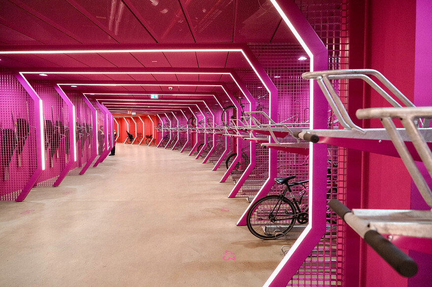 Photos of a futuristic-looking, brightly-lit, colourful bike parking facility and public passage beneath Berlin’s Potsdamer Platz. The red and purple curvilinear space is lined with a variety of vertical and horizontal racks, each separated by a metal grill structure which incorporates LED lighting.