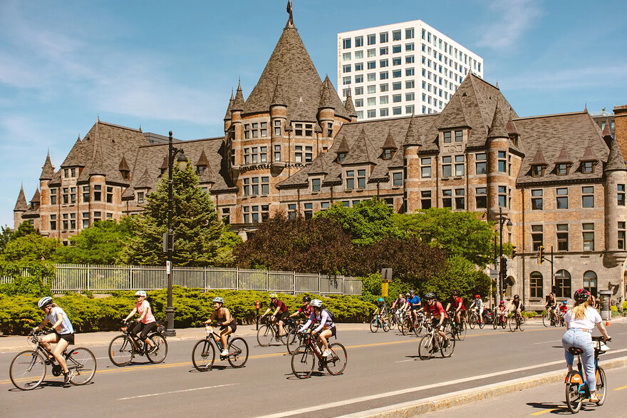 Several dozen bicyclists ride along a wide urban road past a sprawling red-brick building with turrets and a parklike area in front of it
