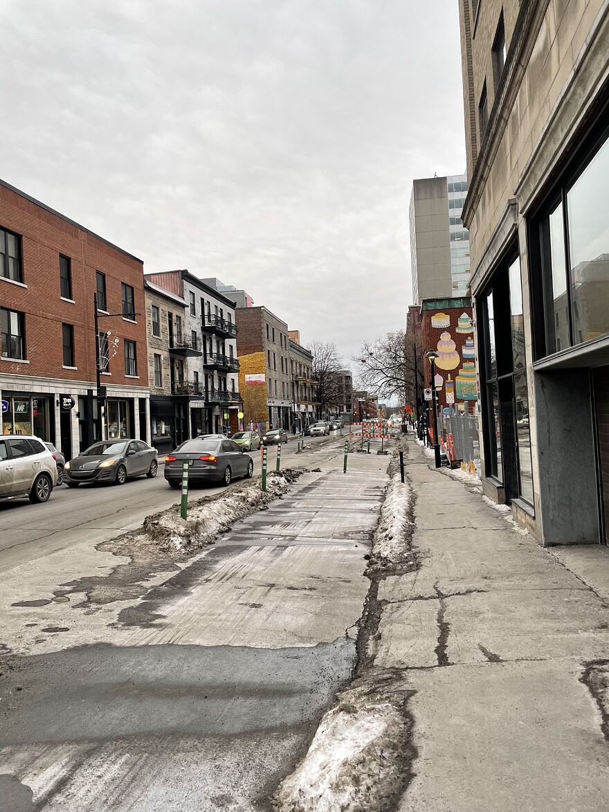 Vue vers le sud sur la rue Atateken. La photo est prise à partir du trottoir ouest, à la hauteur du théâtre l’Olympia. Dans le cadran gauche, on observe la circulation automobile. Une longue file d’automobiles est visible dans la direction nord. Un automobiliste se dirigeant vers le sud s’est arrêté pour naviguer une chicane aménagée à même la piste cyclable. Dans le cadran droit, on observe la piste cyclable Atateken. Au sud du stationnement de l’Olympia, elle dévie vers le centre de la chaussée pour laisser place à un débarcadère, créant par l’effet même, la chicane observée plus tôt.