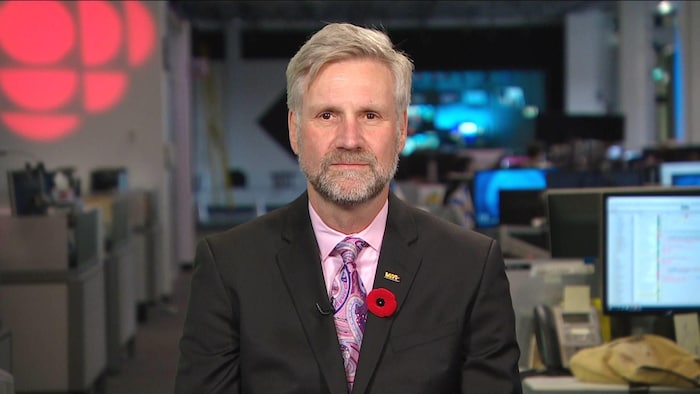 Un homme en complet, assis dans une salle de nouvelles pour une entrevue télévisée, avec le logo de Radio-Canada projeté derrière lui.