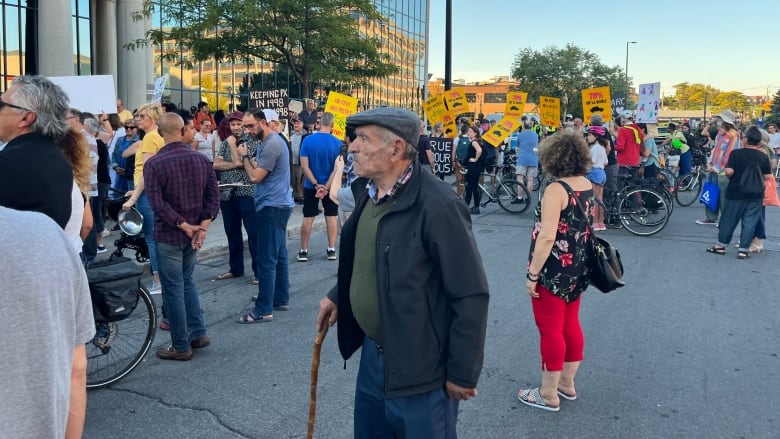 An old man wearing a newsboy cap and a wooden cane stands amid a crowd of protesters carrying signs in favour of bike lanes and against the removal of parking spaces.