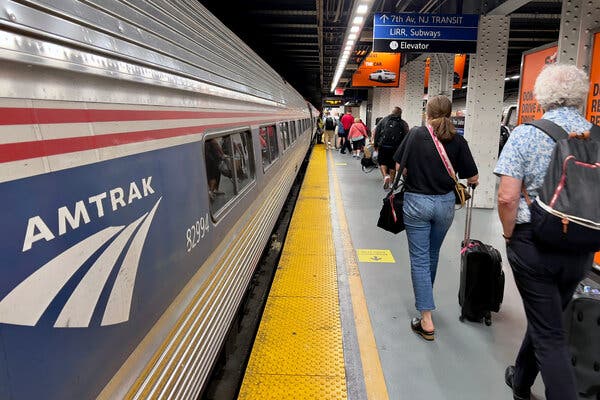 In the left of the frame, an Amtrak train sits at a platform. At the right, people walk with luggage along a yellow caution line down the length of the train.