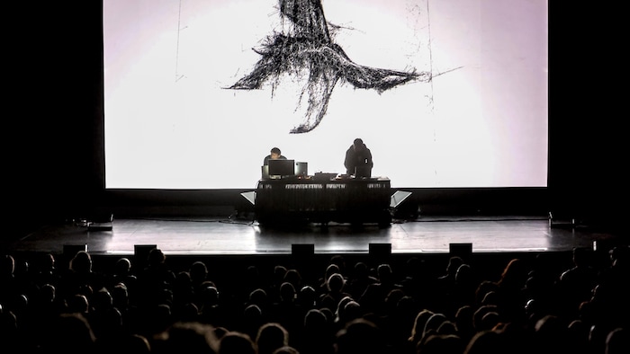 On voit deux artistes sur scène devant un écran blanc présentant une forme organique noire.