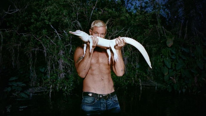 Un homme torse nu tient un petit alligator blanc devant son visage.