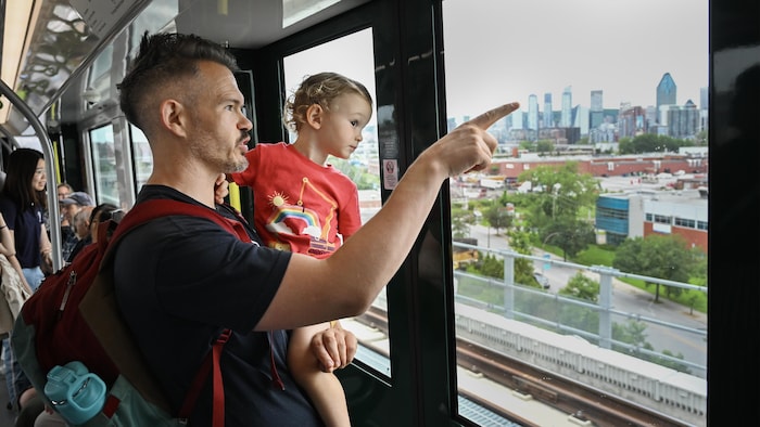 Dans un train, un homme tient son fils dans les bras et lui pointe quelque chose à l'extérieur.