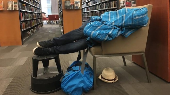Un homme se réfugie dans une bibliothèque publique du centre-ville. Il dort sur un un fauteuil et ses jambes allongées sur un tabouret.