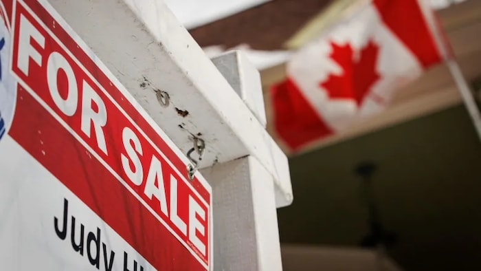 Une pancarte de maison à vendre avec un drapeau du Canada qui flotte au vent sur le terrain en arrière.