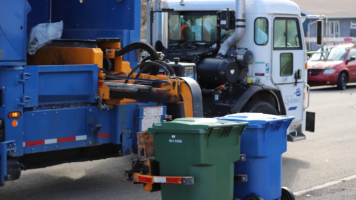 Un camion de collecte des matières résiduelles de la Ville de Québec s’apprête à vider le contenu d’un bac roulant vert dans sa benne à l’aide d’un bras articulé.