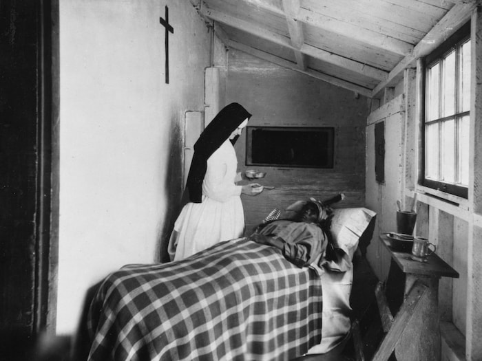 Hospitalière soignant une femme autochtone alitée dans une petite chambre rudimentaire, œuvre du début du 20e siècle.