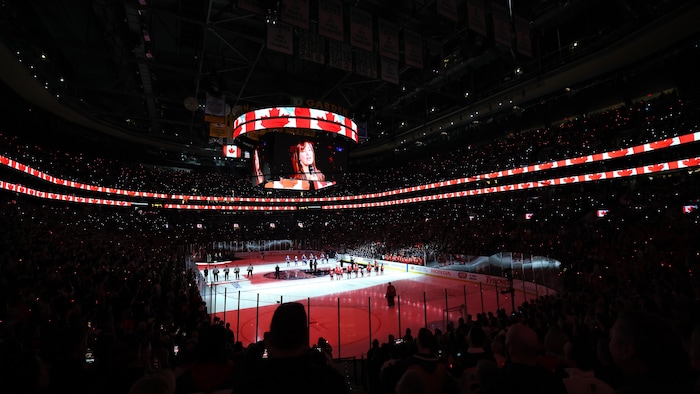 Dans un aréna, un écran au-dessus d'une patinoire retransmet la vidéo d'une femme qui chante un hymne national. On voit aussi des projections du drapeau canadien sur la glace et sur des écrans au-dessus de la foule.