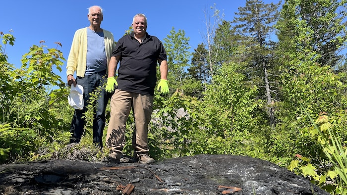 Pierre Racicot et Marcel Poiré sont debout sur un tas de bardeaux d'asphalte jetés en pleine nature.