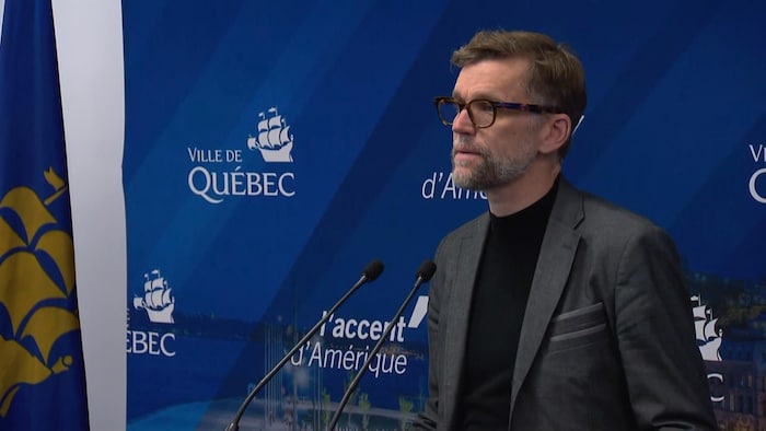 Le maire Marchand parle devant deux micros et un décor affichant le logo de la Ville de Québec.