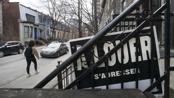 Une jeune femme marche sur un trottoir devant une pancarte de logement à louer posée dans la balustrade d'un escalier.