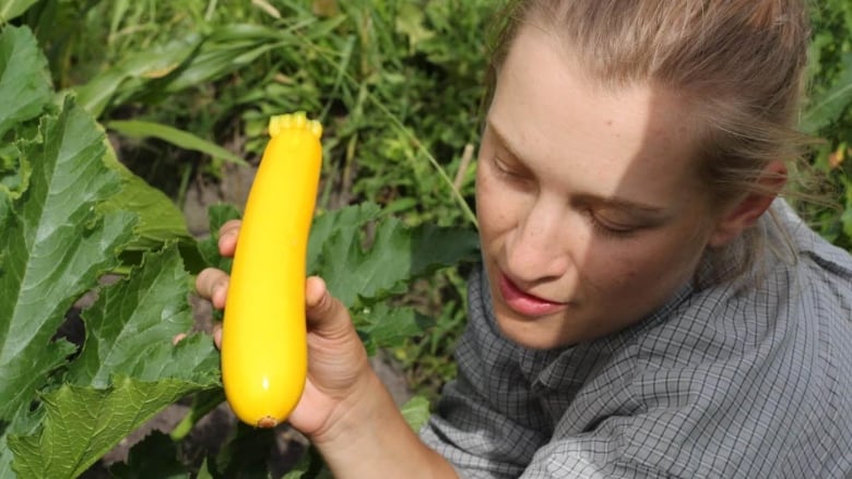 A woman holds up a zucchini in a garden.