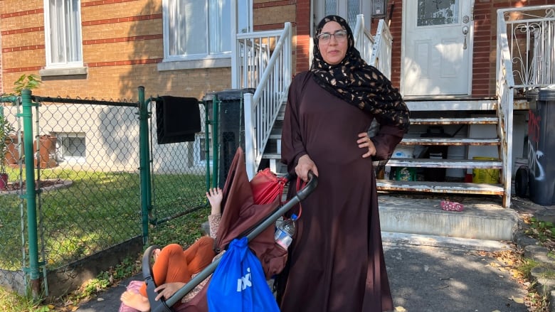 A woman wearing a headscarf and eye glasses stands outside her home with her three-year-old in a stroller.