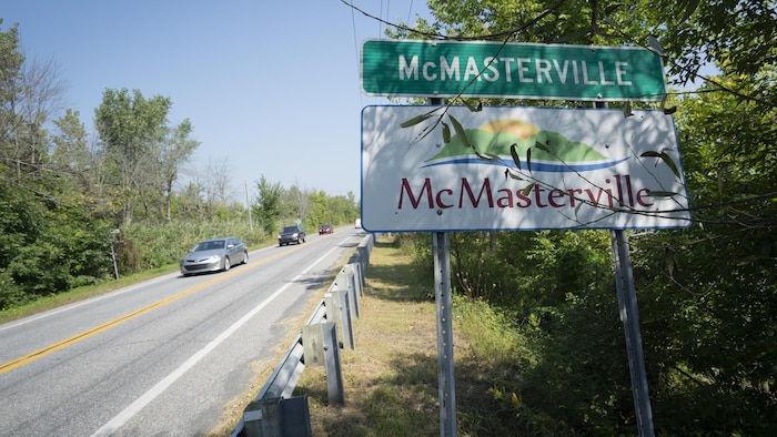 Un panneau annonçant la municipalité de McMasterville en bordure d'une route où l'on voit des voitures.