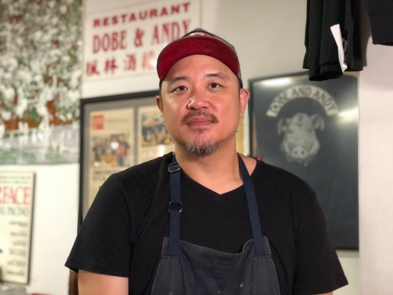 A chef in apron and ball cap looks seriously at the camera. It's a portrait. He's inside his restaurant. Poster and art hang on the wall behind him.