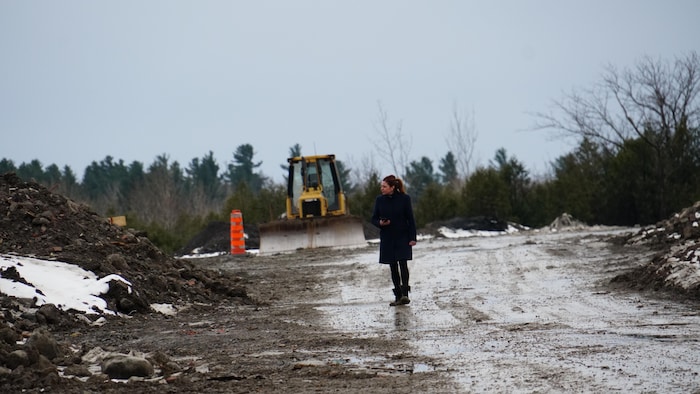Marie-Maude Denis marche sur une route de terre devant une pelle mécanique stationnée et un cône orange.