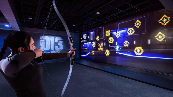 Une personne tient un arc à flèche et pointe vers des tuiles virtuelles au mur.