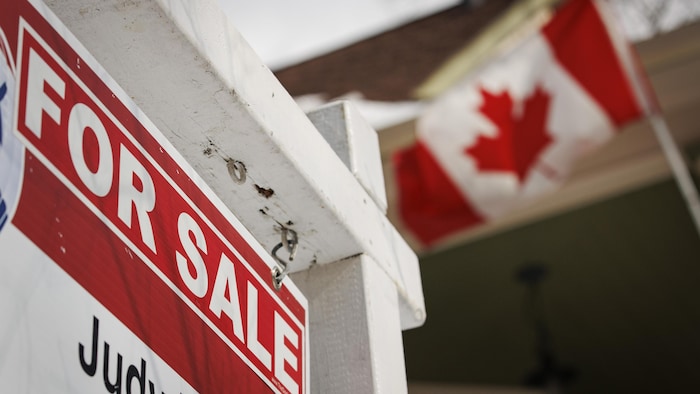 Une pancarte de vente devant une maison qui fait flotter un drapeau canadien.