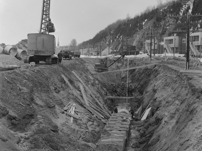 Une pépine et un énorme trou au fond duquel se trouvent deux ouvriers, en hiver, lors du lancement des travaux de construction du boulevard Champlain, en 1962