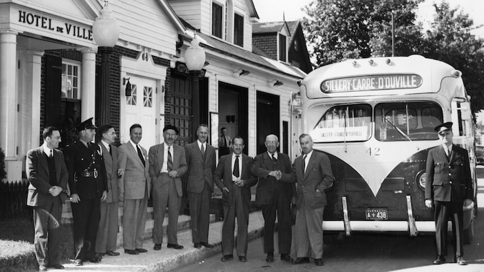 Le maire Gérard Guay et une rangée de dignitaires souriants devant l'ancien hôtel de ville de Sillery lors de l'inauguration de la ligne d'autobus reliant Sillery et Place d'Youville, probablement en 1961.