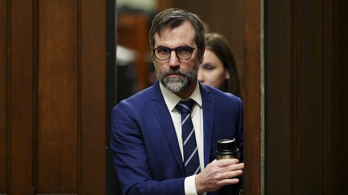 Steven Guilbeault, l'air sérieux, passe le cadre d'une porte. Il tient dans sa main une tasse réutilisable.