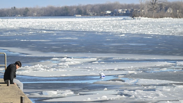 Une personne assise sur le bord d'un quai de béton regarde l'eau à moitié glacée du fleuve.