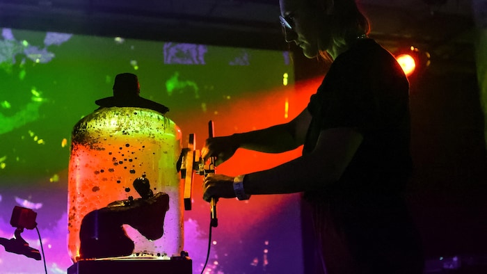 On voit l'artiste devant un bocal avec un fluide magnétique. Il y a des éclairages colorés.