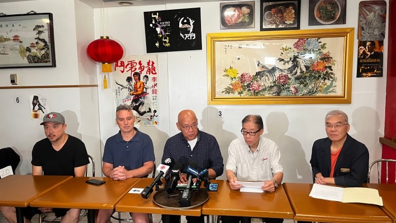 A group of men sit at a table during a news conference. Chinese poster art and a red lantern hang on the wall behind them.