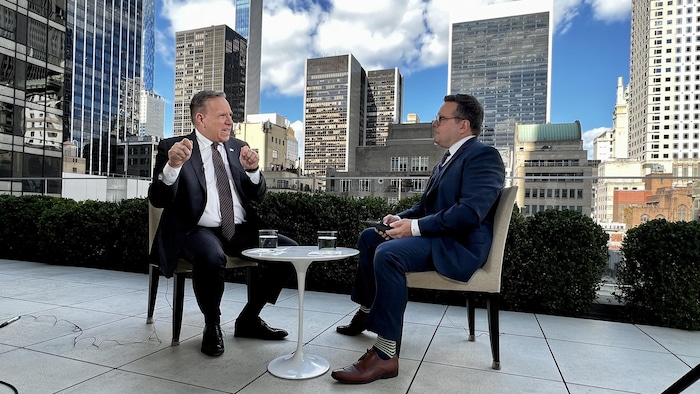François Legault gesticule en discutant avec Daniel Thibeault. Tous deux sont assis devant un panorama de la ville de New York à l'arrière-plan.