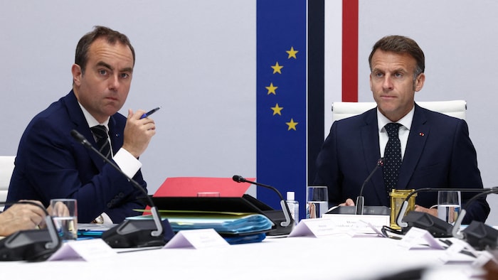 Deux hommes en costume sont assis à une table de réunion, devant le drapeau français et le drapeau européen