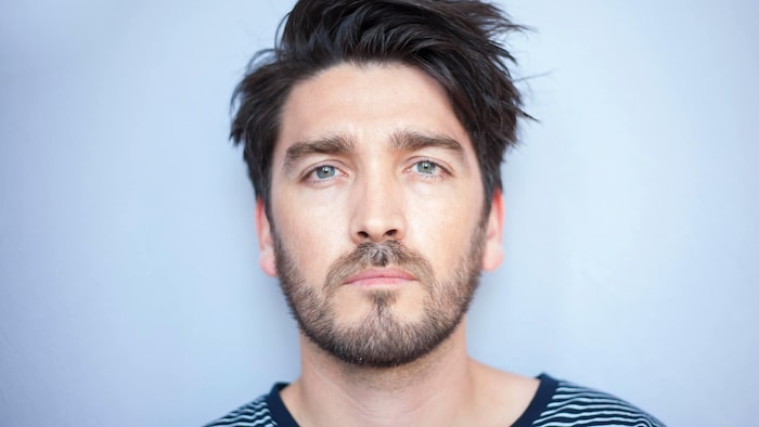 Un homme barbu pose devant un fond blanc et fixe la caméra.