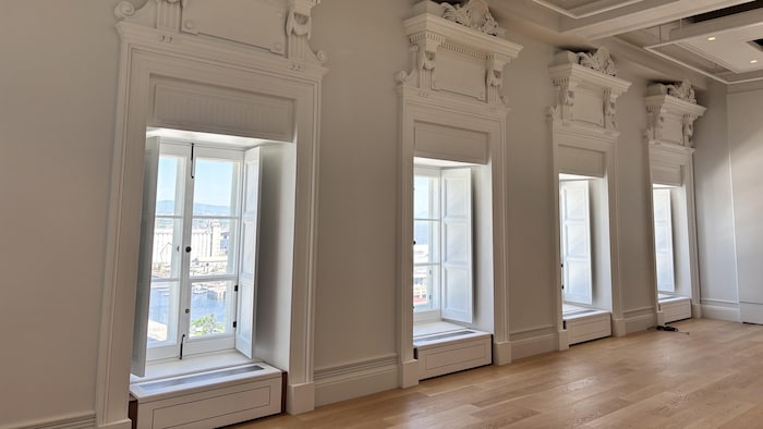 Quatre fenêtres aux moulures sculptées dans une pièce vide, dont la première s'ouvre sur des installations portuaires.