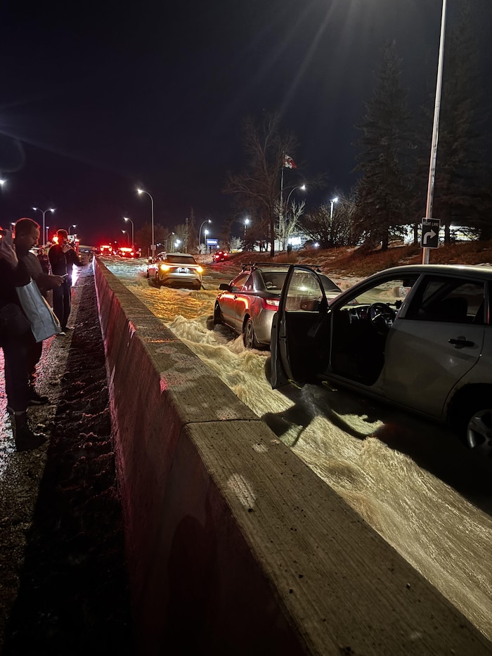 Plusieurs véhicules sont immobilisés dans un important courant d'eau boueuse qui inonde une autoroute la nuit. Une portière de voiture est ouverte au premier plan, tandis que des témoins observent la scène depuis le bord de la route derrière une murette de béton.