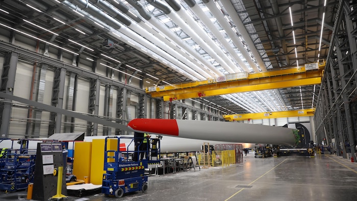 Une grande usine de fabrication avec une longue pale d'éolienne au centre, des ponts roulants jaunes et un monte-charge bleu